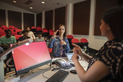 Image of a Lady talking to a presenter in a conference room and other people sitting listening