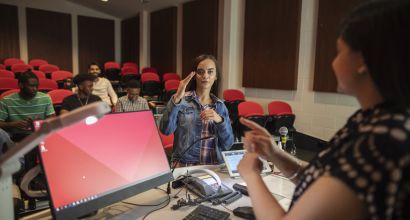 Image of a Lady talking to a presenter in a conference room and other people sitting listening